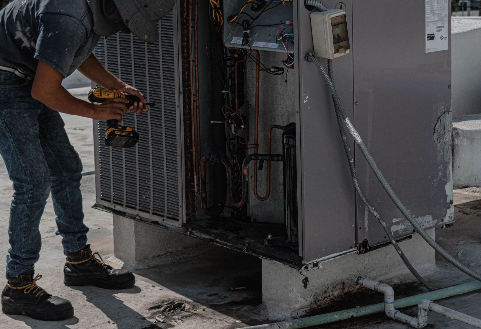 A worker in a bucket hat repairs an outdoor air conditioning unit on a rooftop.